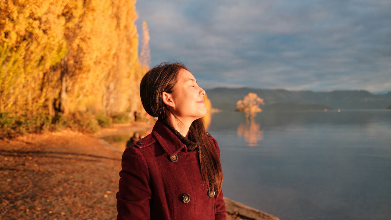 A woman in a red coat stands by the water, gazing at the serene landscape around her.