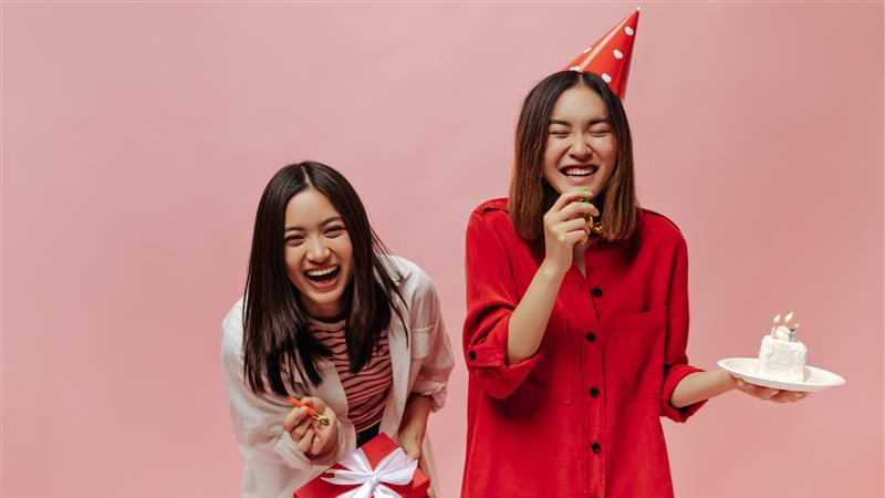 Two women in red party hats smile while holding a cake, celebrating a joyful occasion together. image