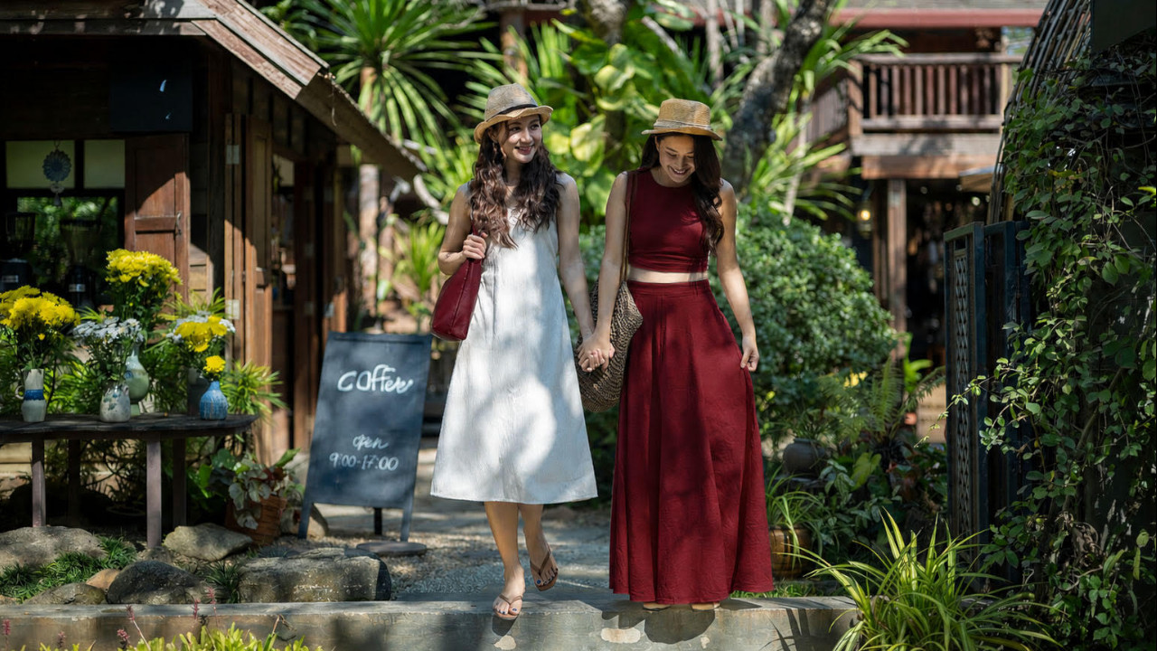 Two women in white dresses and red skirts stroll down a scenic path, enjoying a sunny day together.