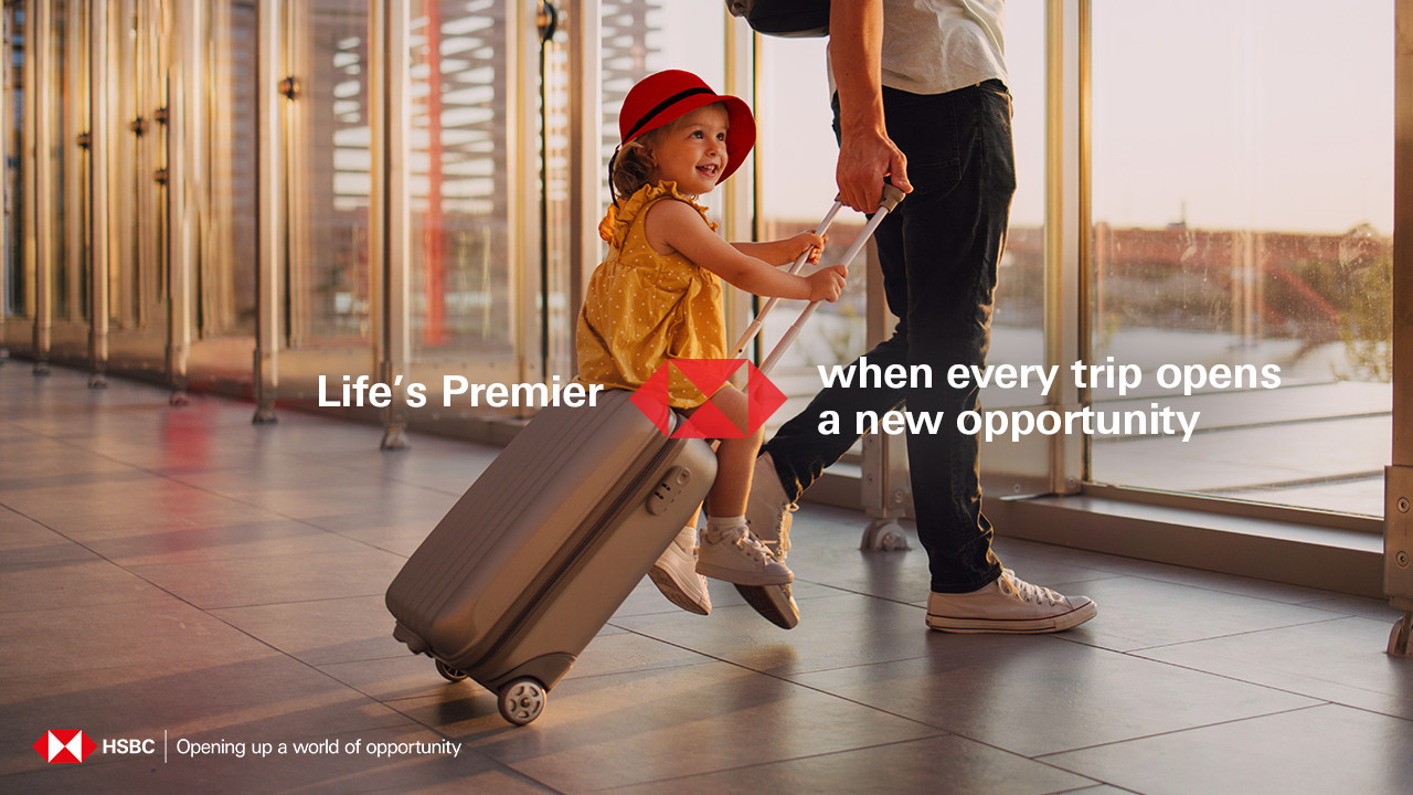 A man pulls luggage with a little girl sitting on top, walking near an airport window.