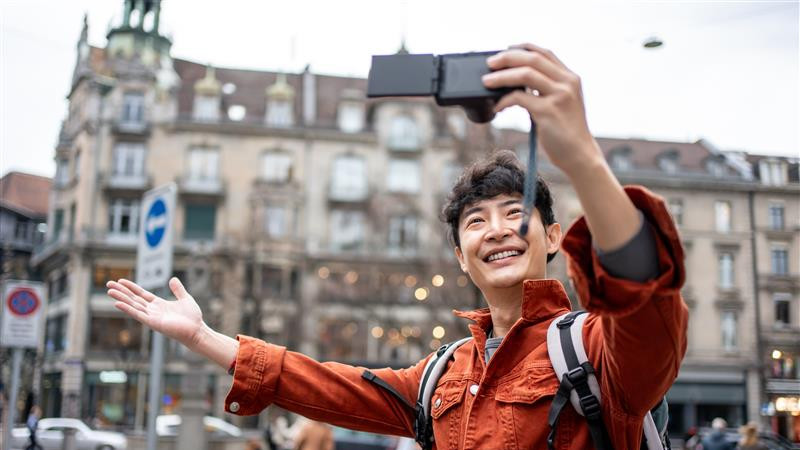 A young man holds his phone at arm's length, smiling as he takes a selfie in a bright, outdoor environment.