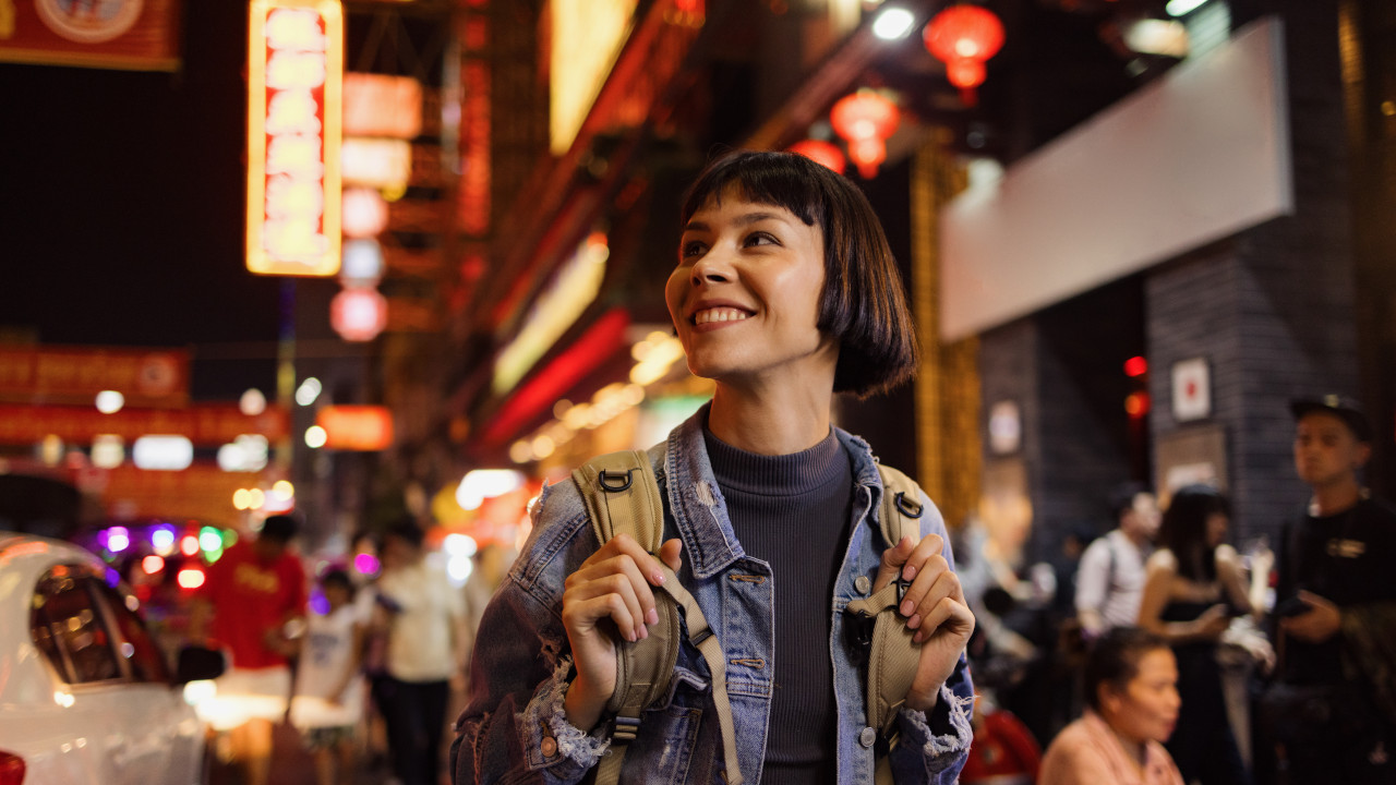 At night, a woman stands on a busy city street, illuminated by neon lights and the glow of passing cars.