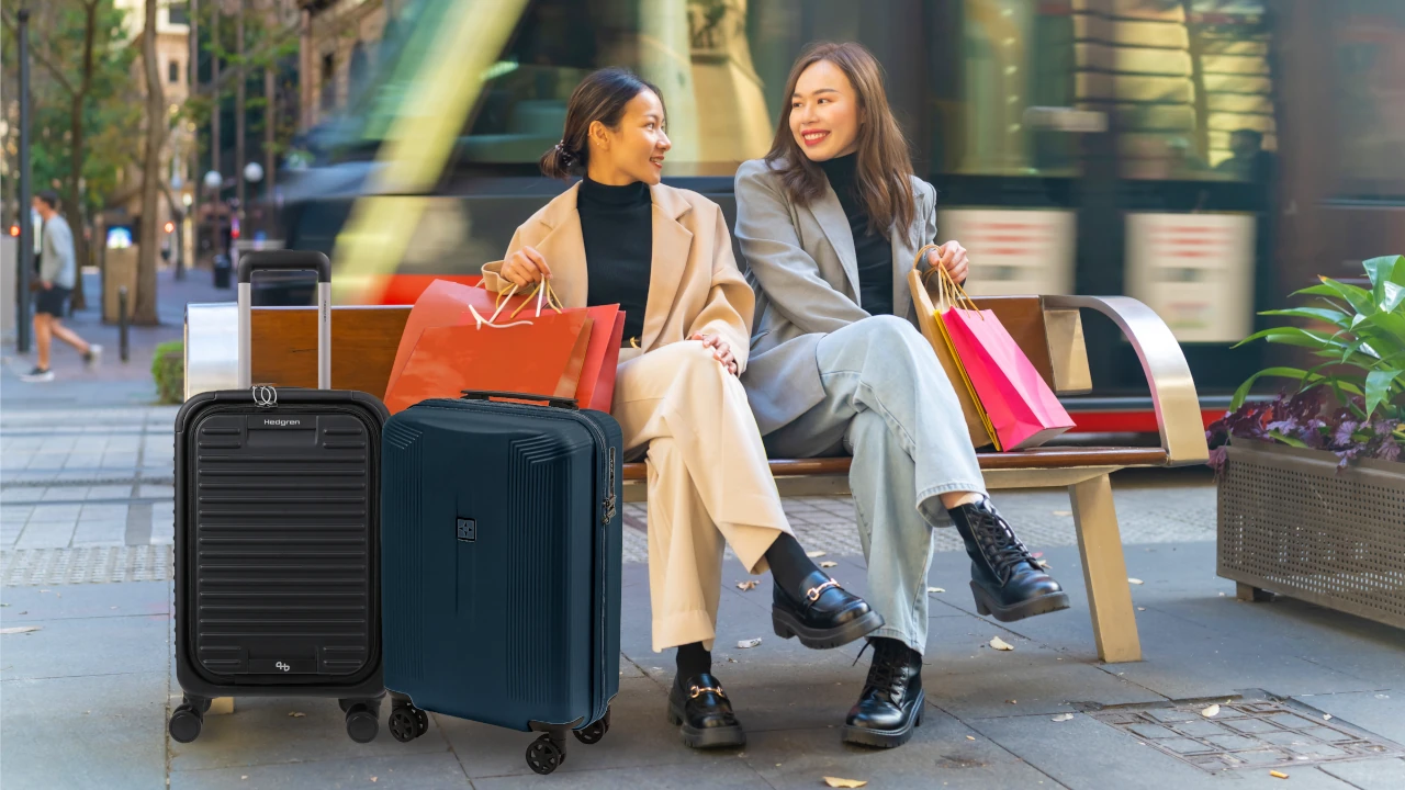 Two women sit on a bench with luggage, engaged in conversation, in a public space.