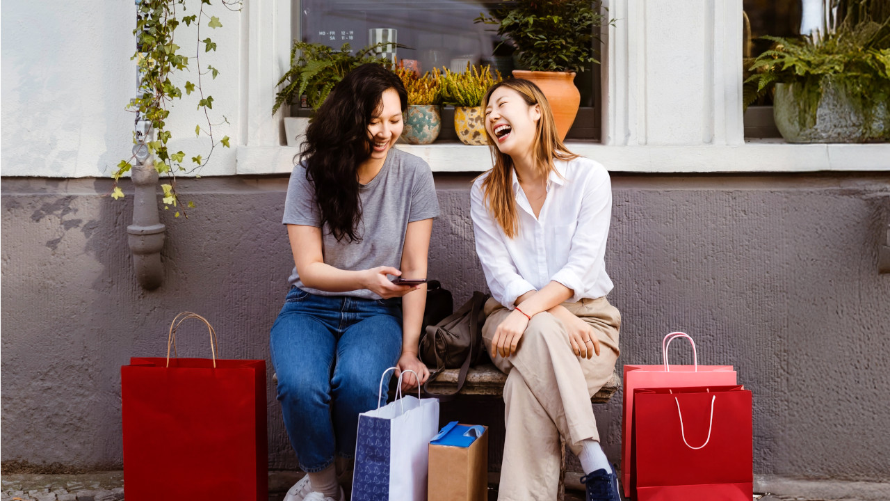 Two women sit on a bench, smiling and holding shopping bags, enjoying a leisurely day out together.